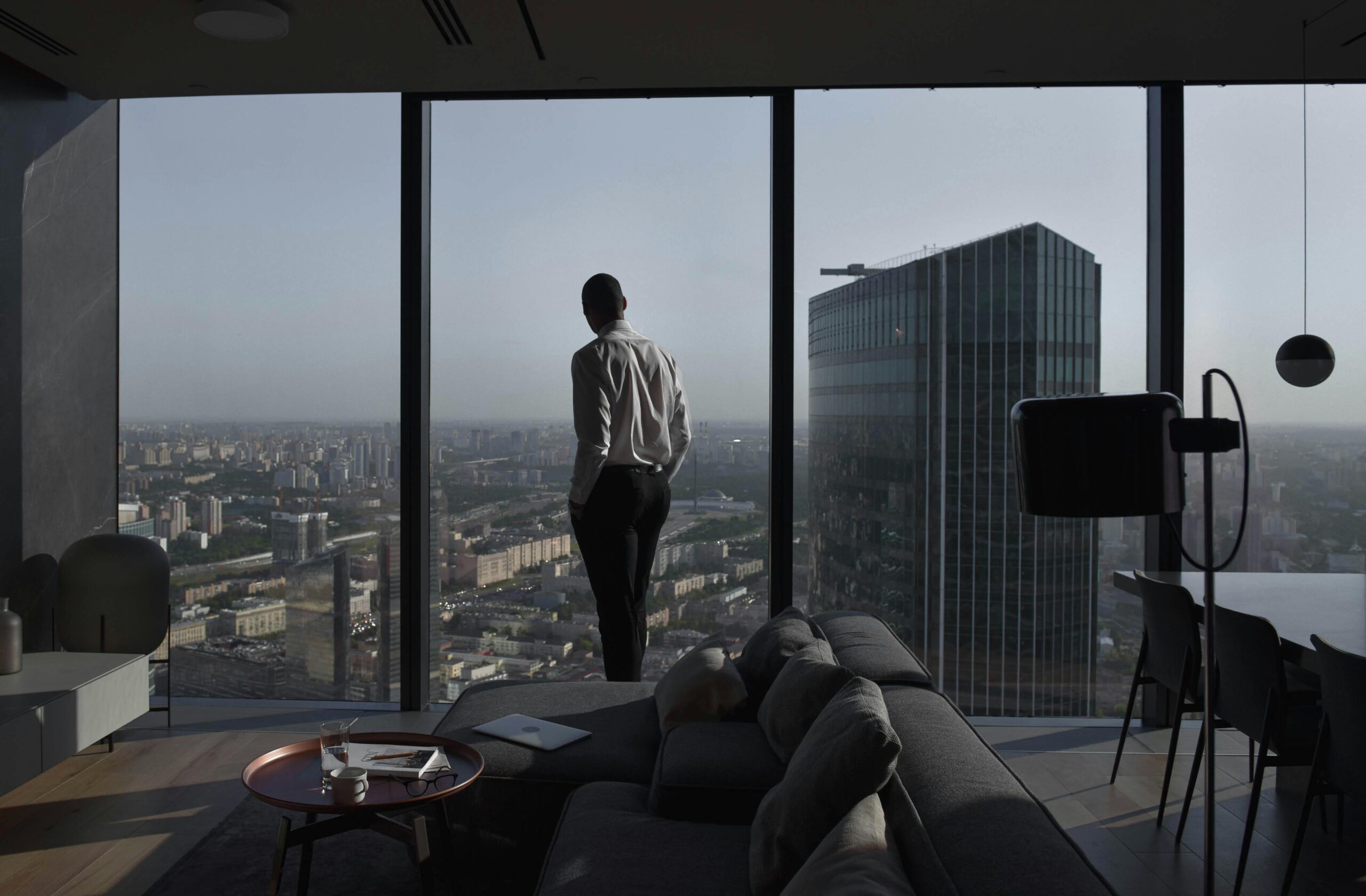 Man in a modern apartment with a panoramic view of an urban skyline and skyscraper.